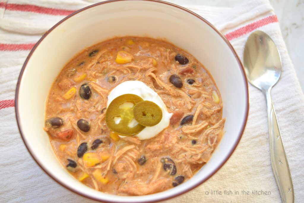 A bowl filled with auburn colored cream cheese chicken chili sits inside a white bowl with a brown rim. Corn, black beans and bits of tomato are visible in the soup. A spoon lays beside the bowl. 