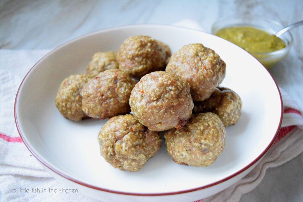 Pesto Turkey Meatballs are ready to serves. They are in a white bowl on top of a white tea towel with a red strip. A small bowl of green pesto sits on the side. 