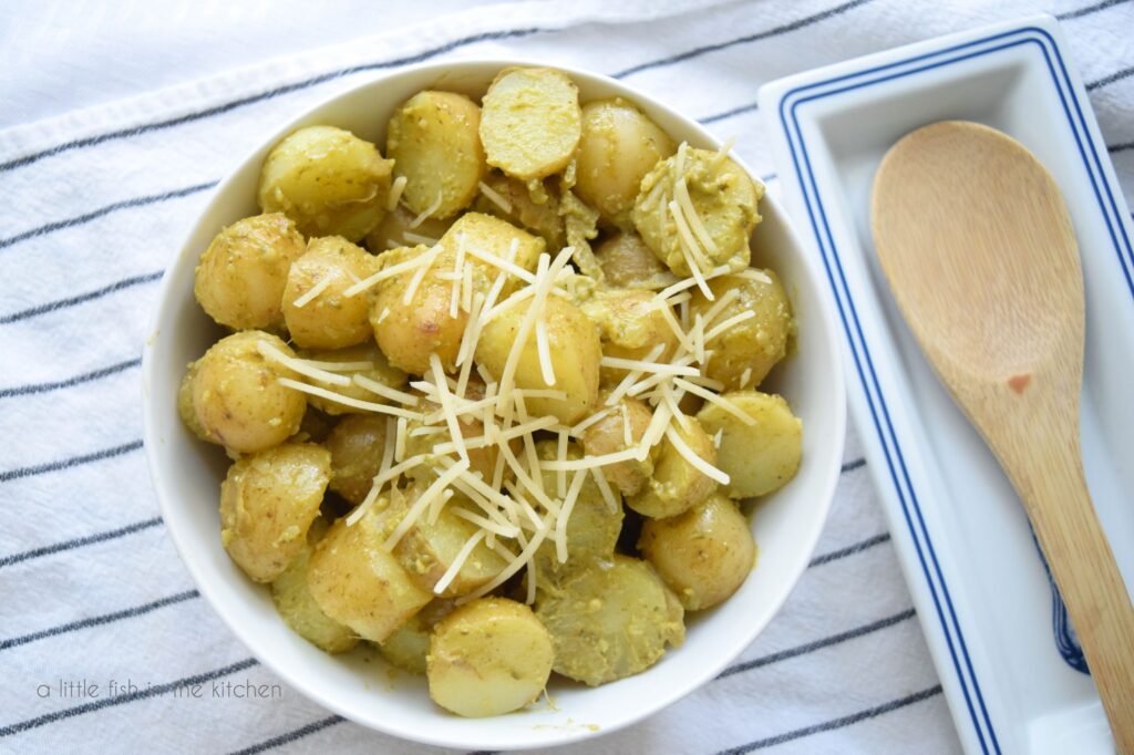 A white bowl filled with Pesto Potato Salad sits on top of a white towel with black strips. The salad is topped with shredded Parmesan cheese. A spoon site beside the bowl in t a blue and white spoon rest. 