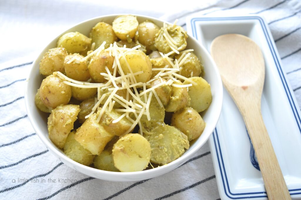 A white bowl filled with Pesto Potato Salad sits on top of a white towel with black strips. The salad is topped with shredded Parmesan cheese. A spoon site beside the bowl in t a blue and white spoon rest. 