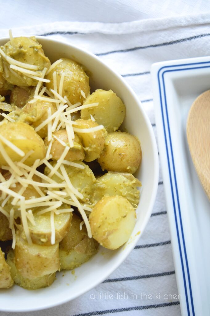 A white bowl filled with Pesto Potato Salad sits on top of a white towel with black strips. One of of the bowl is featured in this image. The salad is topped with shredded Parmesan cheese. A spoon site beside the bowl in t a blue and white spoon rest. 