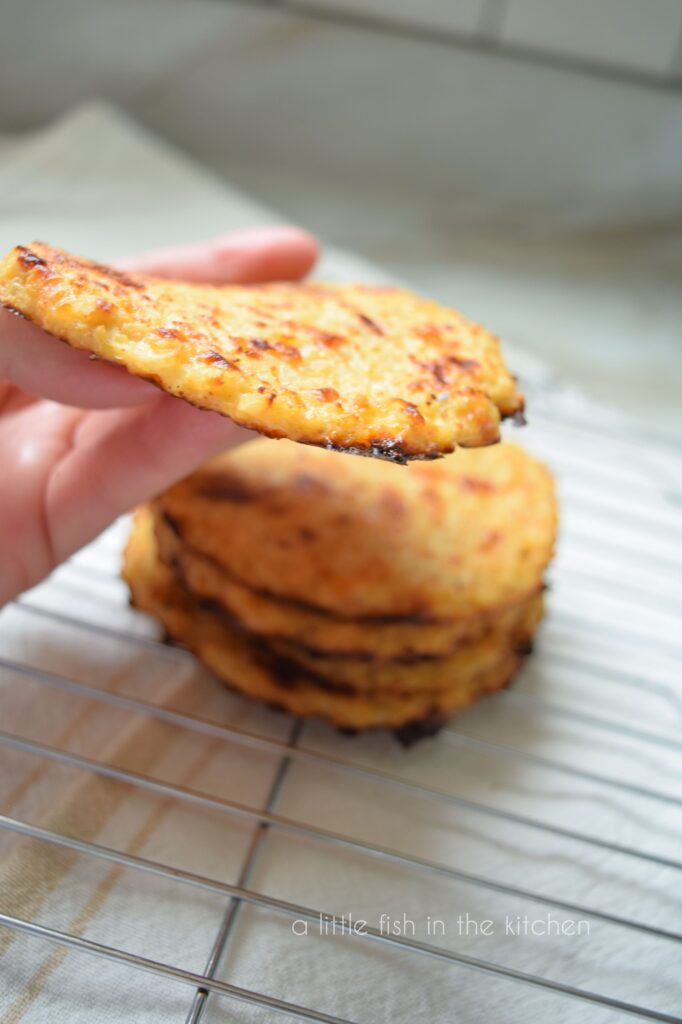 A hand is holding one sandwich thin so the side can be seen. A stack of 5 homemade cauliflower sandwich thins is slightly blurred behind it on a cooling rack.