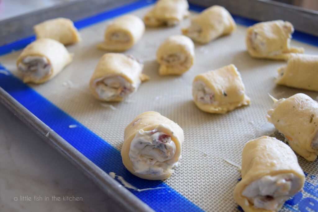Unbaked chive, onion and bacon roll ups rest on a baking sheet and are ready to go into the oven. 