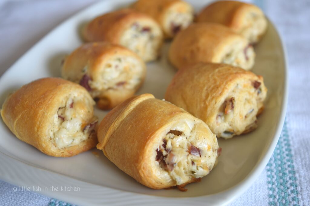 Eight cheesy chive, onion and bacon roll ups sit on a serving dish. The pastry is golden brown and the cheese filling is showing from the sides. 