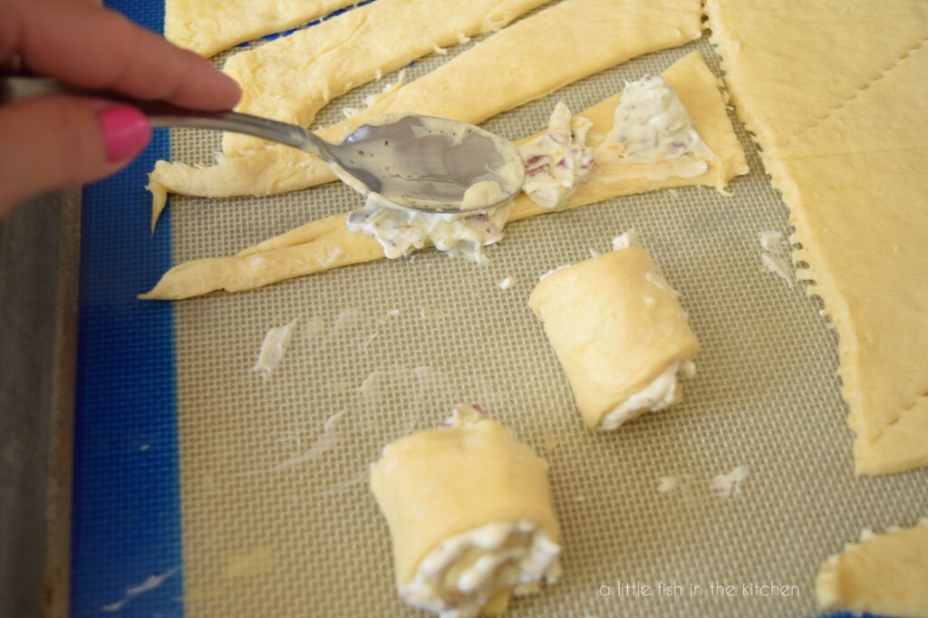 The back of a spoon is used to spread the cream cheese mixture onto strips of crescent roll dough.