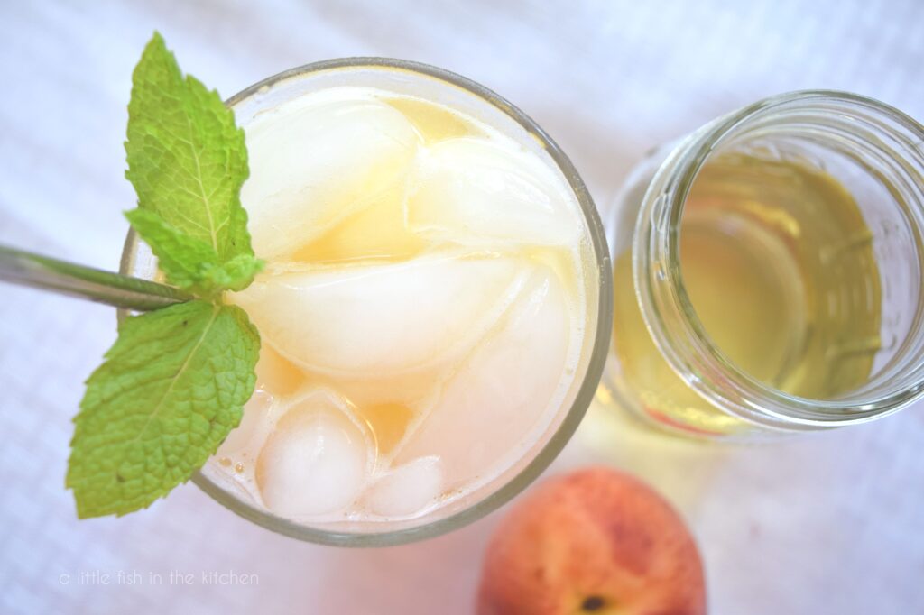 The top of a glass full of Minty Peach Cooler is shown garnished with fresh mint. A jar of syrup sits beside the drink. 