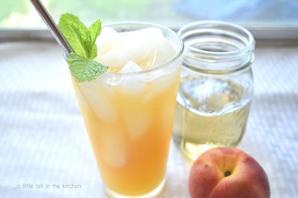 A peach coolered drink is in a clear glass with ice cubes and garnished with a sprig of fresh mint. A whole fresh peach sits beside the glass. A mason jar filled with mint simple syrup sits slitght blurred behind the beverage. 