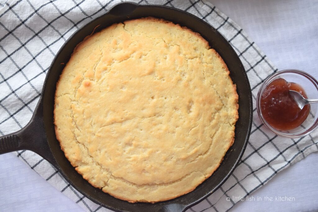 Golden quick bread sits in a black cast iron skillet. A small bowl with strawberry jam and a spoon sits beside the skillet on a black and white checkered tea towel. 