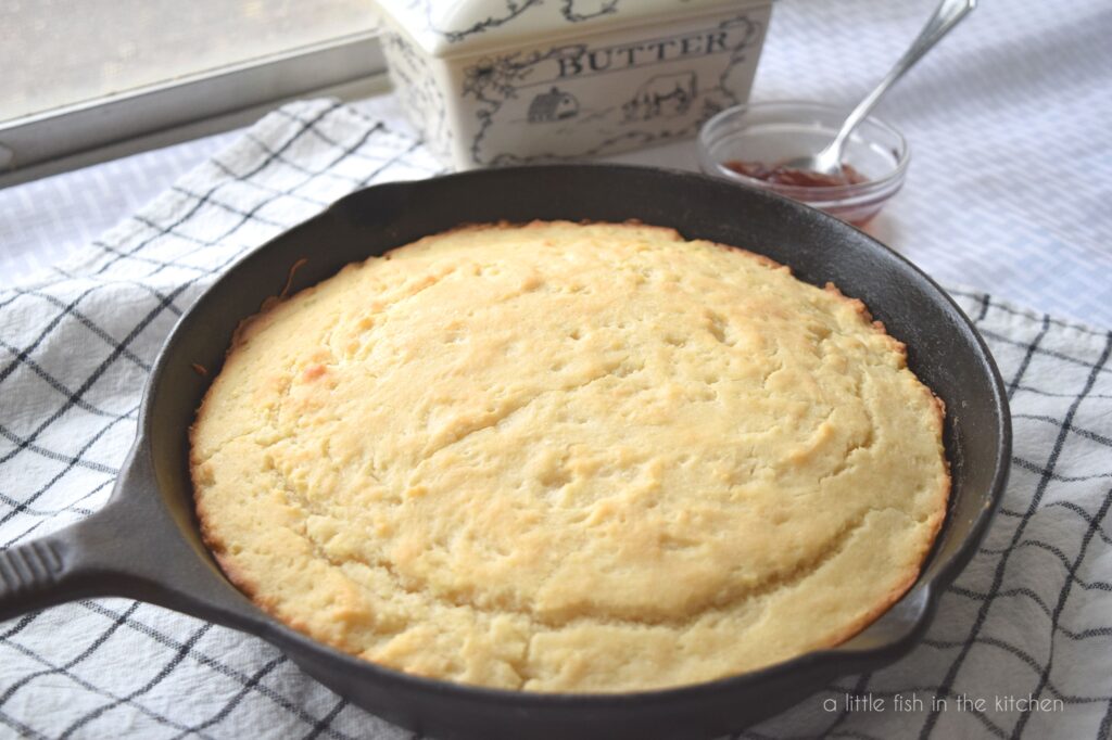 Golden bread sits in a black cast iron skillet. The skillet sits on a black and white checkered tea towel. A butter dish and a small bowl with strawberry jam sits beside the skillet. 