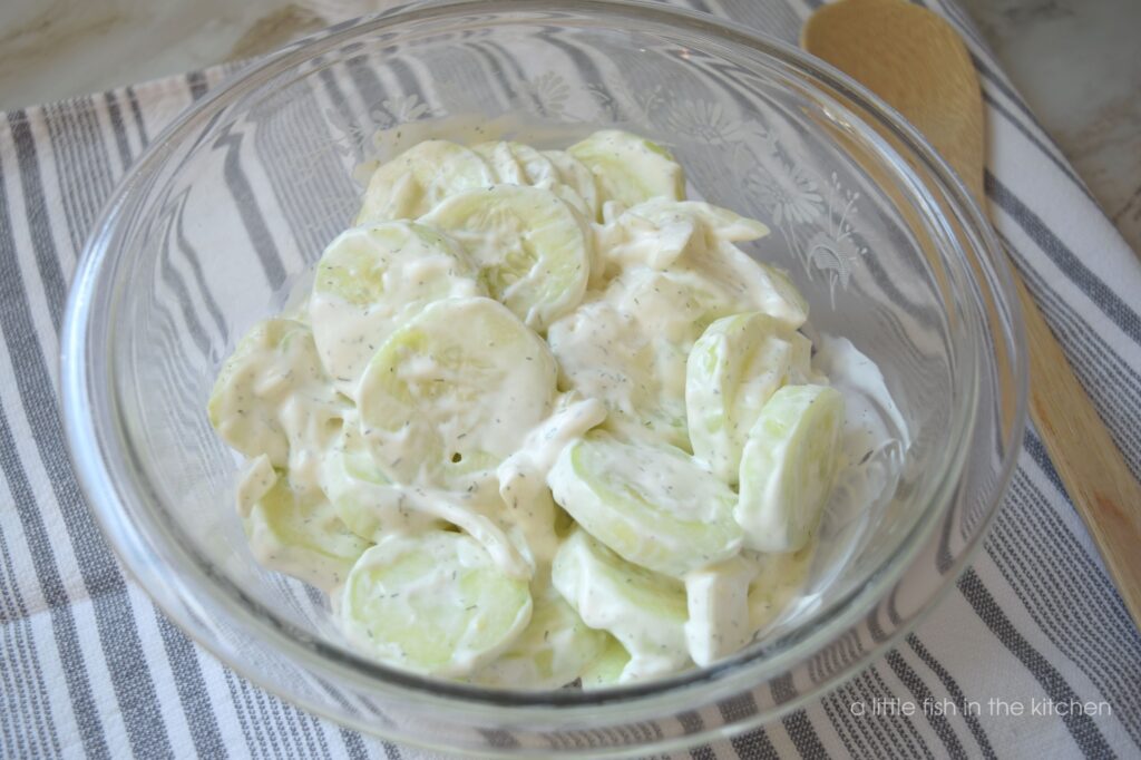 Slices of fresh cucumber are coated in creamy sour cream. This salad is in a clear bowl resting on a gray and white tea towel. A wooden spoon rests beside the glass bowl for serving. 