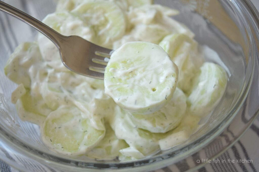 A fork is holding a sour cream-coated cucumber slice from a bowl of creamy cucumber salad. The large bowl filled with salad is slighy blurred in the background. 
