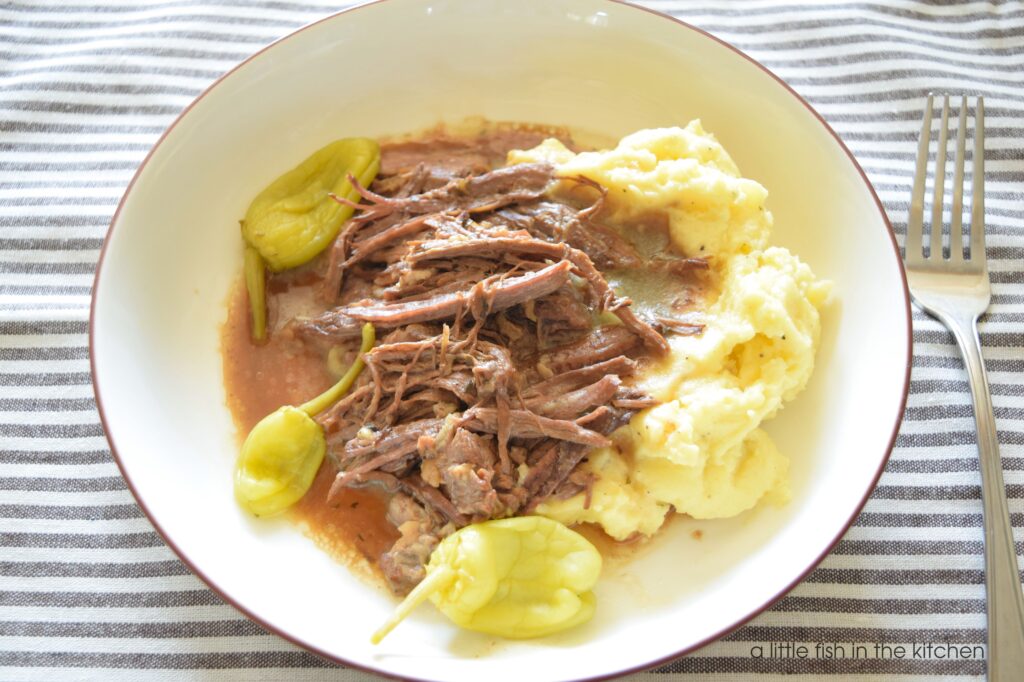 A slightly angled view of a white bowl filled with shredded Mississippi pot roast on a bed of mashed potatoes and gravy. 