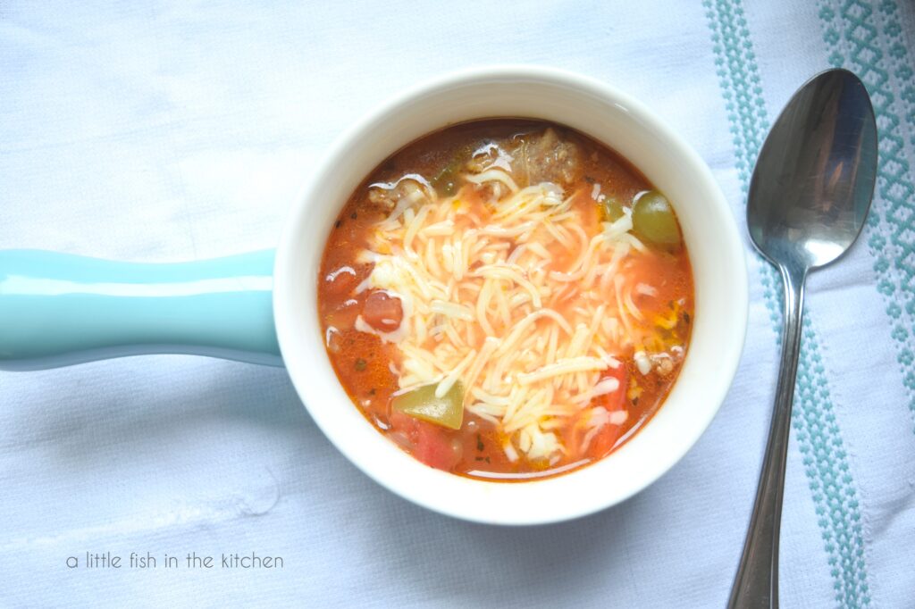 Italian sausage and pepper soup in a light blue soup bowl. It's topped with melted mozzarella shreds. A spoon is beside the bowl. 