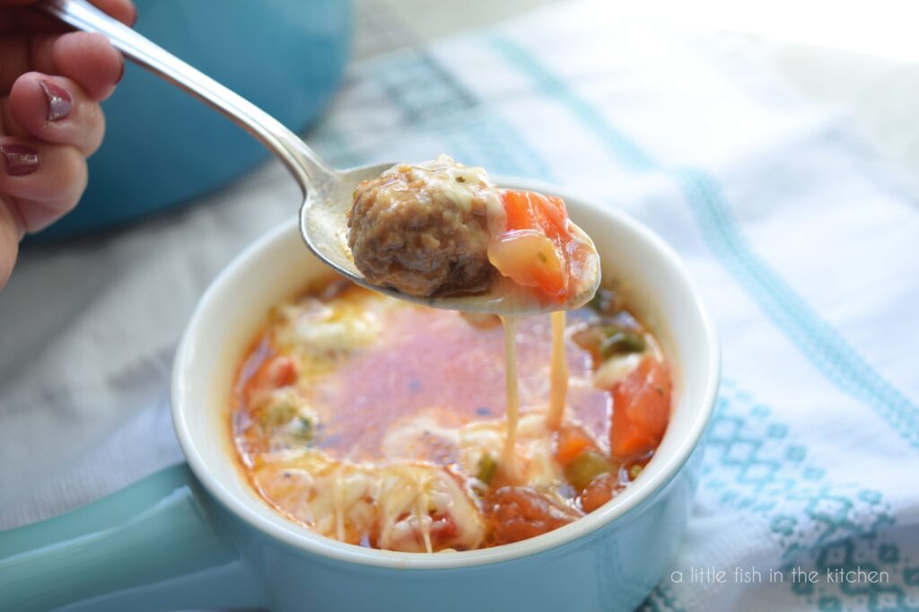 A spoon loaded with sausage and tomatoes is hovering over a bowl of soup. These is melted cheese dangling from the spoon. 