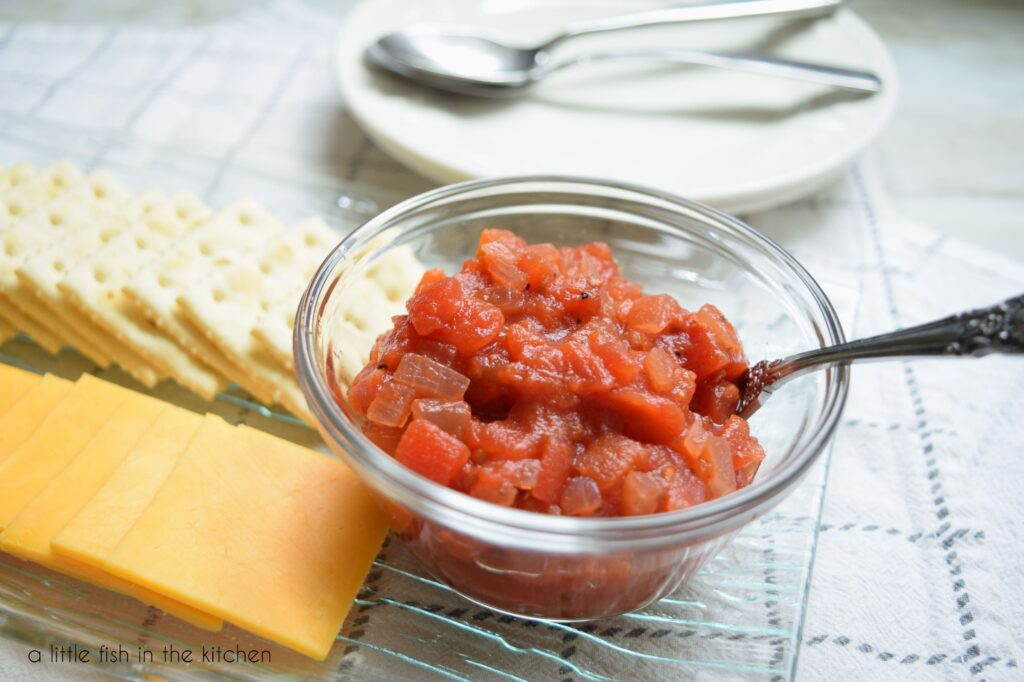 A small clear glass bowl is filled with tomato chutney. Bits of tomato,  cooked red onion and black pepper are visible in a close up picture. A spoon is nestled in the bowl. The bowl sits on a rectangular glass plate that is filled with a row of each yellow cheese slices and saltine crackers. 