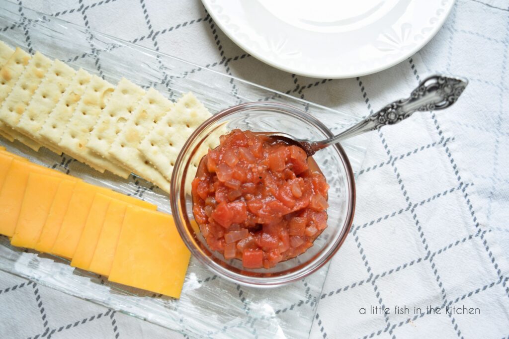 A small clear glass bowl is filled with dark red tomato chutney. This picture is taken directly above the bowl and cooked diced Bits of tomato, cooked red onion and black pepper are visible. A spoon is nestled in the bowl. The bowl sits on a rectangular glass plate that is filled with a row of each yellow cheese slices and saltine crackers. The plate sit son a white and gray striped tea towel. 