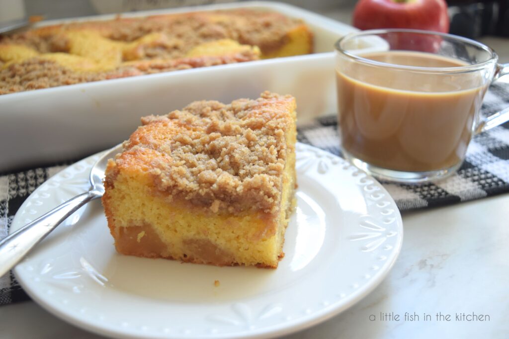 A slice of apple pie cake is on a white cake plate. The layers of crunchy crumb topping, buttery yellow cake and bits of apple pie filling are well-defined in this slice of cake. A fresh cup of coffee in a clear coffee mug rest just beside the plate. 