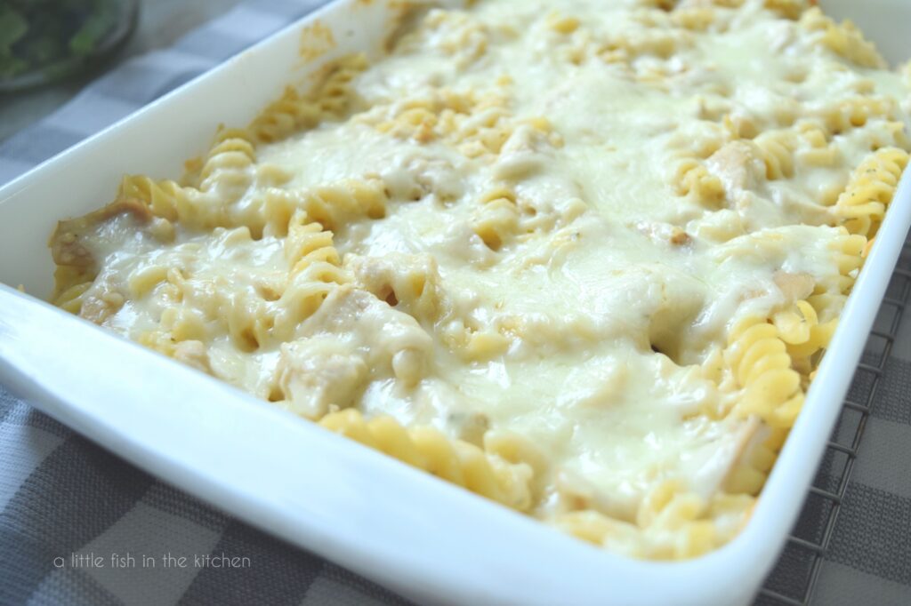 The Easy Alfredo Bake Casserole dish is shown from the top. It is a closer shot of the pasta, chicken and cheese seen in the casserole. It is in a white casserole dish and sits on a grey and white checkered tea towel.  