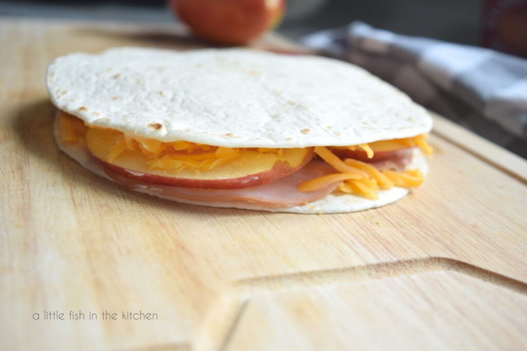 An ungrilled quesadilla sits on a wooden cutting board. Slices of ham, apple and shredded cheese are visible on the sandwich's edge. 