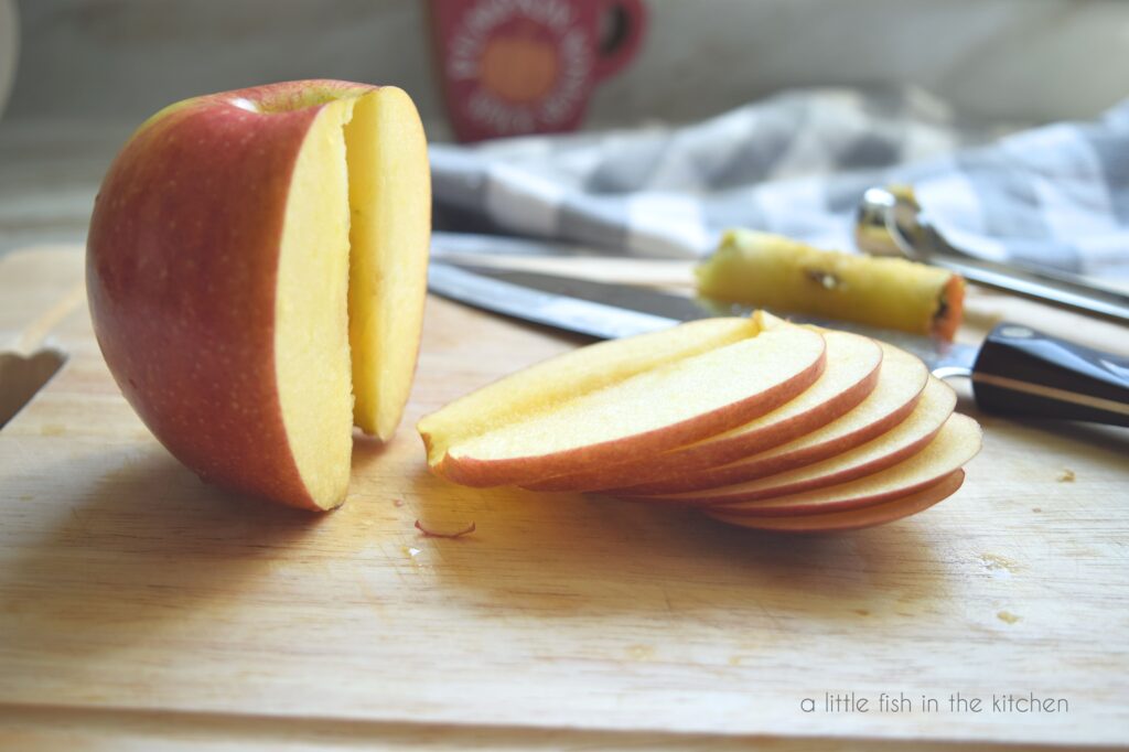 A red apple sits on a wooden cutting board. The apple is sliced in half. One half is whole and the other half is in six thin slices. 