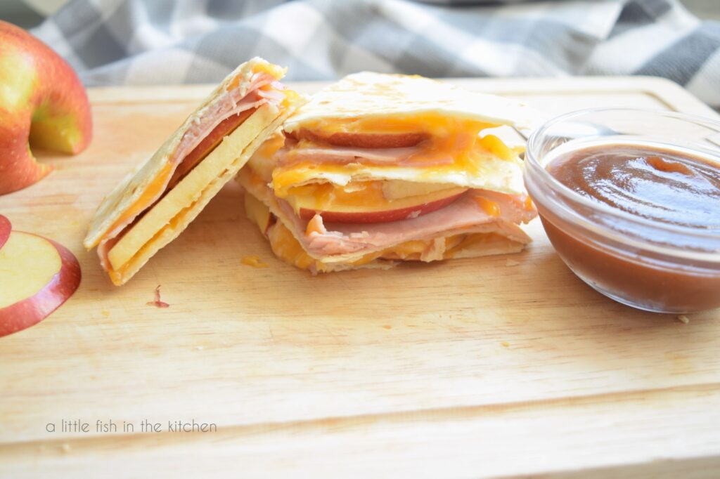 A ham, apple and cheddar quesadilla is sliced into 4 pieces. Three pieces are stacked upon each other, and one is leaning upright against the stack. A small bowl of apple butter sits on the cutting board to the right. 