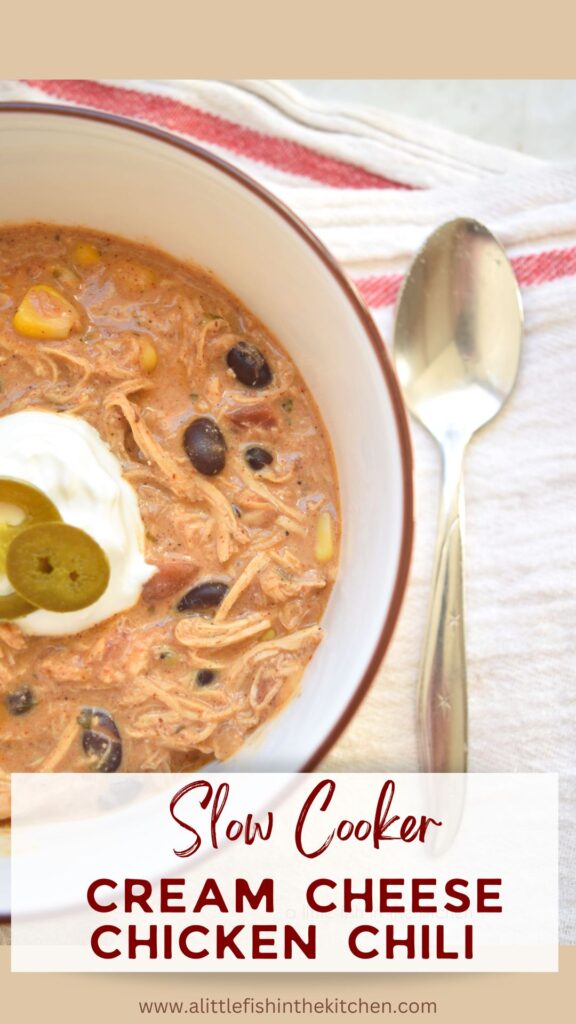 Pin image for this recipe. A bowl filled with auburn colored cream cheese chicken chili sits inside a white bowl with a brown rim. Corn, black beans and bits of tomato are visible in the soup. A spoon lays beside the bowl. 
