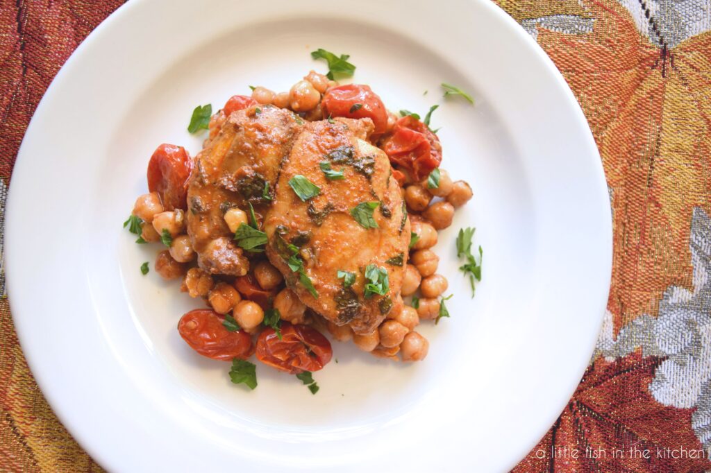 A roasted chicken thigh sits on a bed of roasted chickpeas and tomatoes on a white plate. The entire portion is coated in an amber-colored sauce and is garnished with chopped, fresh parsley. 