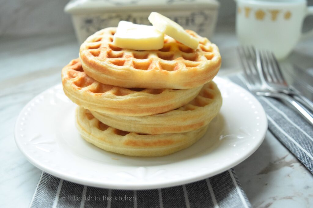A stack of 4 small, golden waffles sits on a white plate. Two pats of butter rest on top of the waffle stack. A butter crock, a small pitcher and two forks are slightly blurred in the background. 