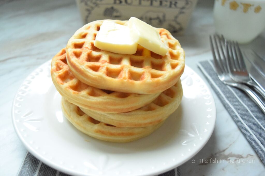 A stack of 4 small, golden waffles sits on a white plate. Two pats of butter rest on top of the waffle stack. A butter crock, a small pitcher and two forks are slightly blurred in the background. This picture focuses  more on the top of the waffle stack. 
