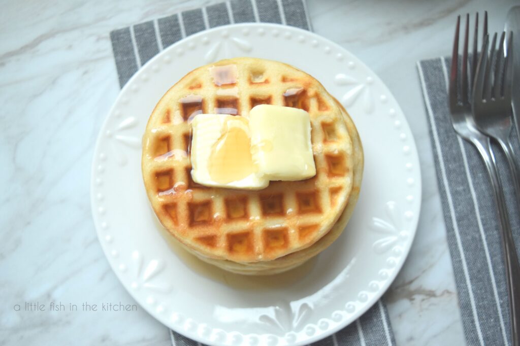 A small stack of waffles sits on a white plate. This pictures focuses on the top waffle in the stack. it's covered with maple syrup and two pats of butter. Two forks sit beside the plate on top of a grey cloth napkins with white stripes. 