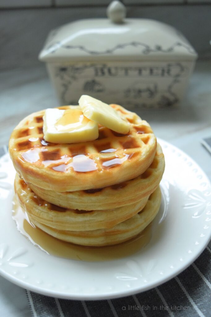 A stack of 4 small, golden waffles sits on a white plate. Two pats of butter rest on top of the waffle stack and the waffles are covered in glistening maple syrup.  A butter crock, a small pitcher and two forks are slightly blurred in the background. This picture focuses  more on the top of the waffle stack. 