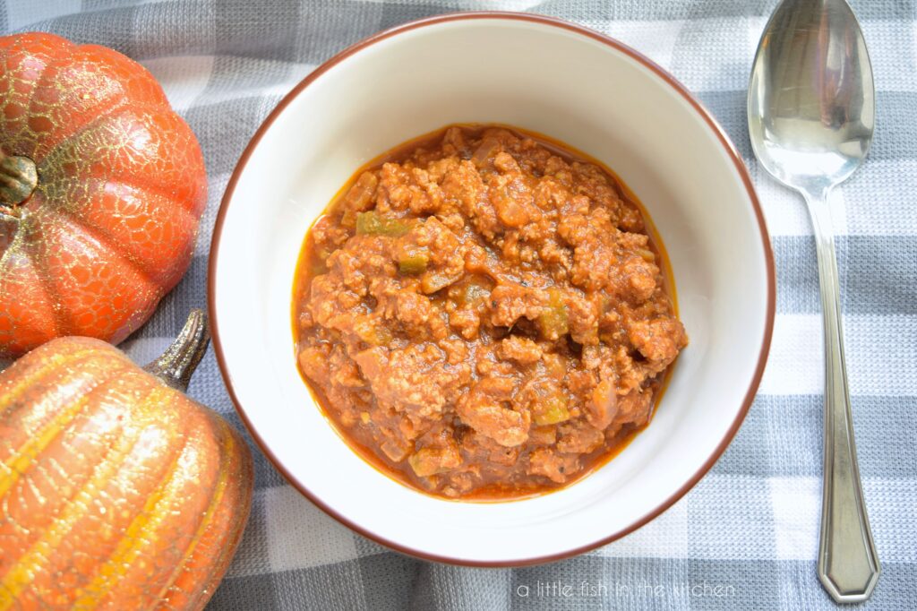 A white bowl with a brown rim is filled with pumpkin chili. Two small decorative pumpkins sit beside the bowl on top of a gray and white checkered kitchen towel. 