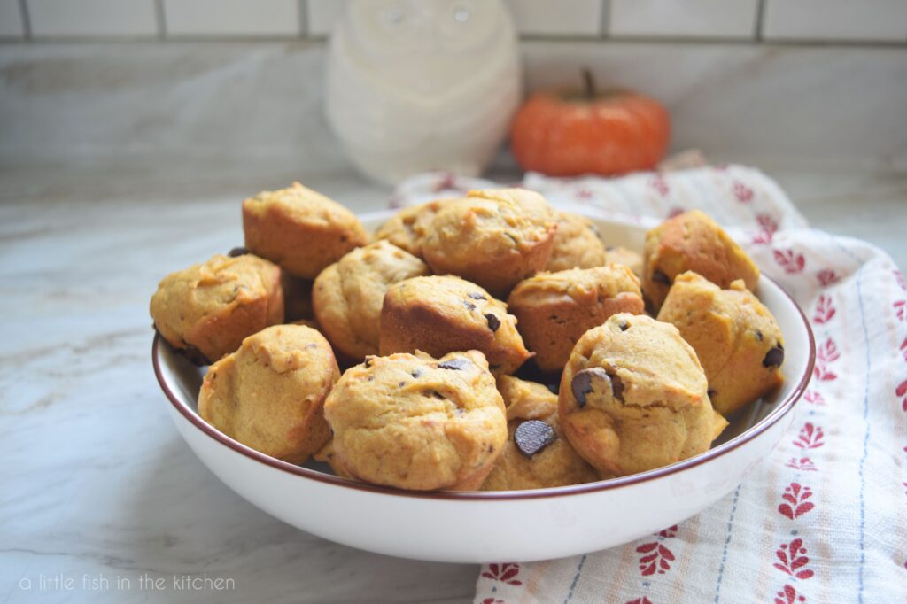 Mini Pumpkin Chocolate Chip Muffins have an earthy orange tint and are speckled with chocolate chops. They are in a white serving bowl, a decorative orange pumpkin is slightly blurred in the background.