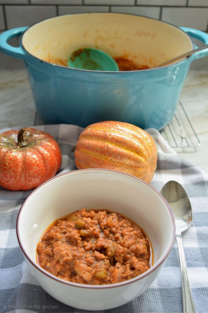 A white bowl filled with pumpkin chili is is featured. There is a spoon beside the bowl as two small decorative pumpkins behind the bowl. A teal dutch oven with a ladle inside is in the background. 