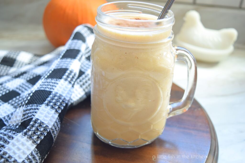 A light orange-colored Pumpkin Maple Protein Smoothie is in a mason jar with a metal straw, ready to drink. A black and white checkered kitchen towel, a small pumpkin and a white ceramic rooster are in the back drop. 