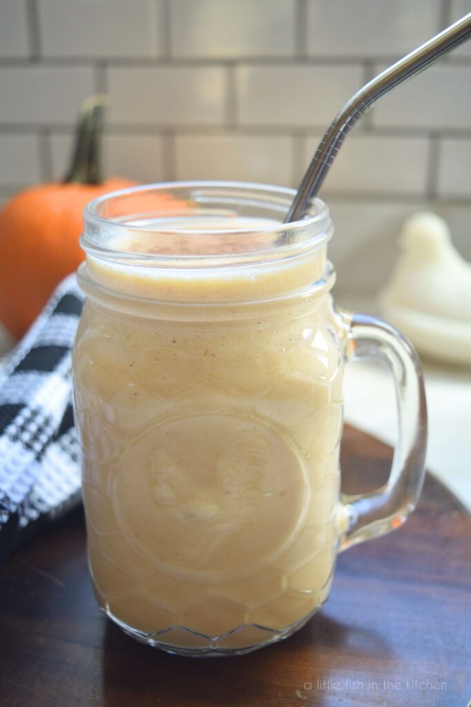 Pumpkin Maple Protein Smoothie is in a mason jar with a metal straw, ready to drink. A black and white checkered kitchen towel, a small pumpkin and a white ceramic rooster are in the back drop. 