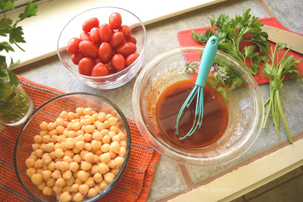 The amber-colored marinade sits in a small, clear mixing bowl. It's surrounded by other small bowls filled with fresh, cherry tomatoes, fresh parsley sprigs and whole chickpeas. 