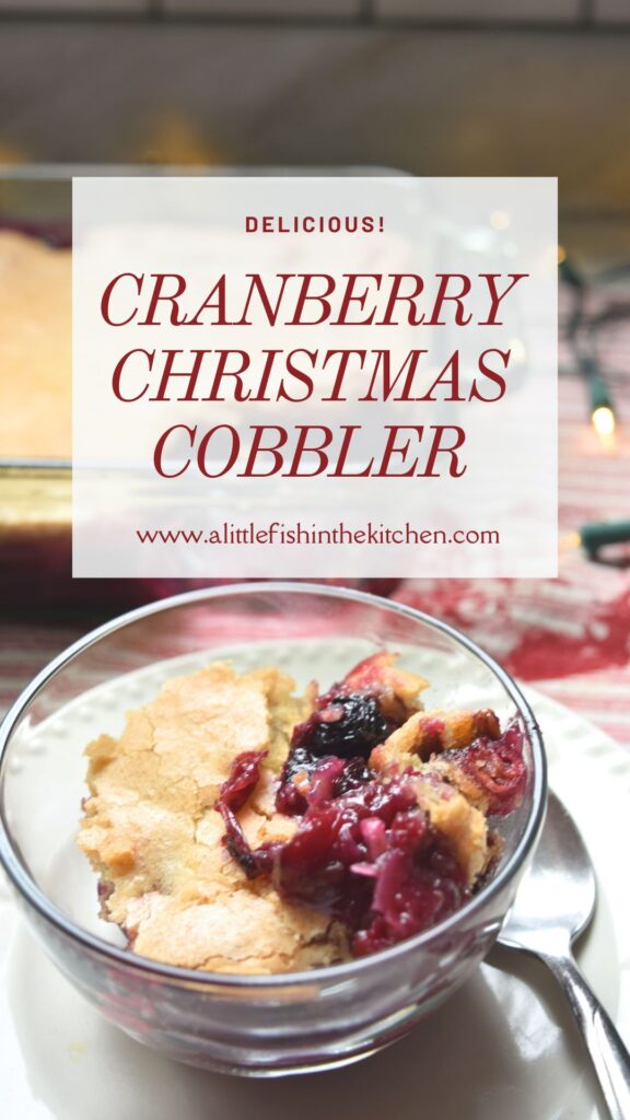 Pin image of this recipe. A clear glass bowl contains a serving of cranberry cobbler. The picture is a close up of one serving of cobbler. The deep red fruit filling is seen on top. Bits of cranberries and blueberries are visible in it. The glass bowl is on top of a small white plate, a spoon is resting beside it. The golden crust is visible on top and some of the deep red cranberry filling can be seen also. The whole casserole and clear baking dish, a red and white striped tea towel and white Christmas lights are slightly blurred in the background. A white overlay is on top of the photo with the recipe and website titles typed on top in burgandy font.