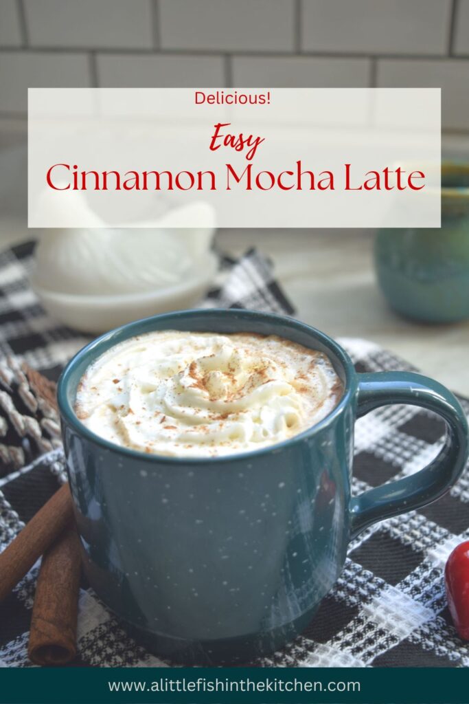 This is a Pin image for Cinnamon Mocha Latte. The name of the recipe and website in contained in a white overlay with the words in red font. A close up shot in a portrait style photo of a dark teal coffee mug  filled with Cinnamon Mocha Latte and topped with swirl of whipped cream sits atop a black and white checkered tea towel. Two cinnamon sticks and a red, heart-shaped ornament sit beside the mug. A decorative ceramic, chicken and a small, teal ceramic mug are slightly blurred in the background against a white subway tile wall. 