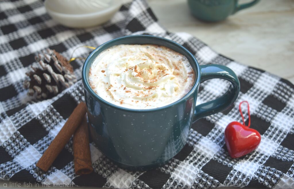 A dark teal coffee mug is filled with Cinnamon Mocha Latte and topped with swirl of whipped cream sits atop a black and white checkered tea towel. Two cinnamon sticks and a red, heart-shaped ornament sit beside the mug. A decorative ceramic, chicken and a small, teal ceramic mug are slightly blurred in the background.  