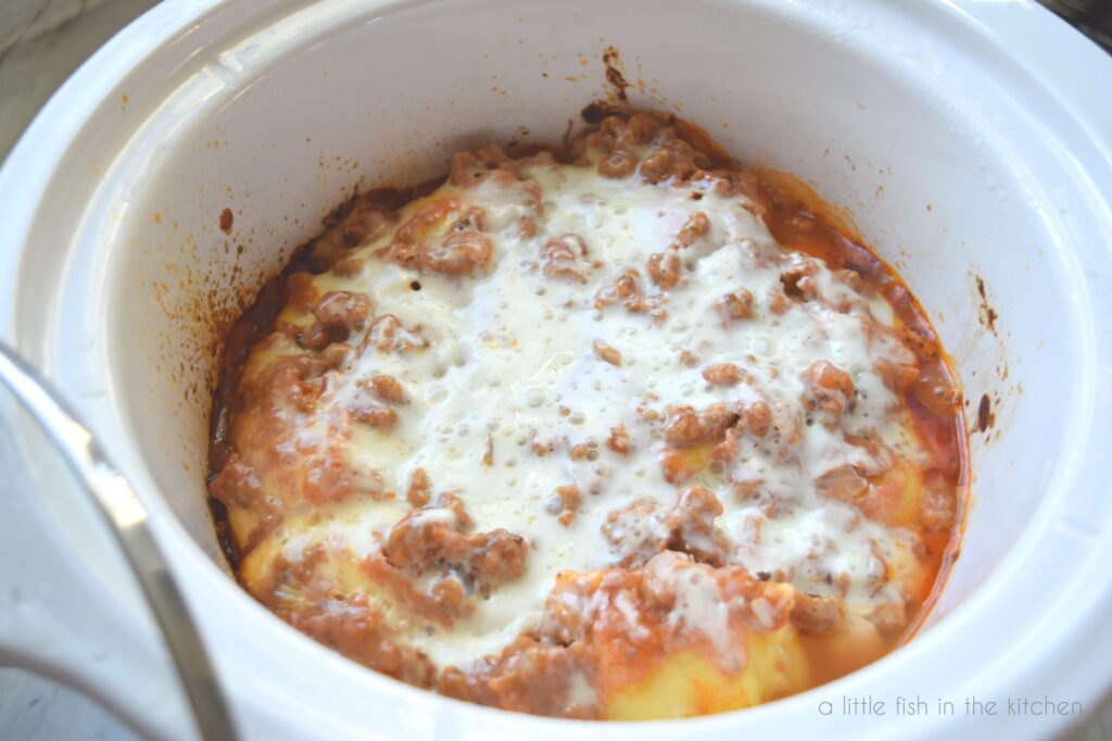 A ravioli lasagna sit in the crock of a white slow cooker crock. Melted cheese and bits of ground Italian sausage are visible on the top. A pool of marinara sauce is on the sides. 