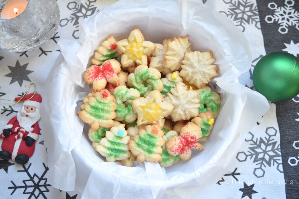A holiday tin lined with parchement paper is filled with petite spriz cookies. The cookies are shaped like Christmas trees, snow flakes, wreaths and bows. They're topped with decorative sugars that is either red, yellow or green. The tin sits on a white tea towel that is printed with gray snowflakes. Two small Christmas ornaments lay beside the tin for decoration. A large glass tea light holder shaped like a snowball is visible in the background. 