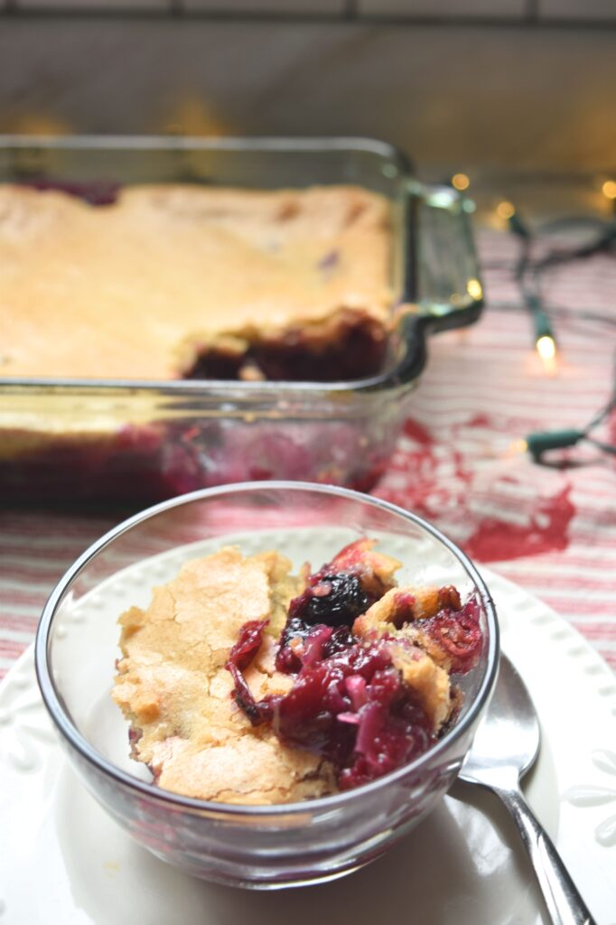 A clear glass bowl contains a serving of cranberry cobbler. The picture is a close up of one serving of cobbler. The deep red fruit filling is seen on top. Bits of cranberries and blueberries are visible in it. The glass bowl is on top of a small white plate, a spoon is resting beside it. The golden crust is visible on top and some of the deep red cranberry filling can be seen also. The whole casserole and clear baking dish, a red and white striped tea towel and white Christmas lights are slightly blurred in the background.