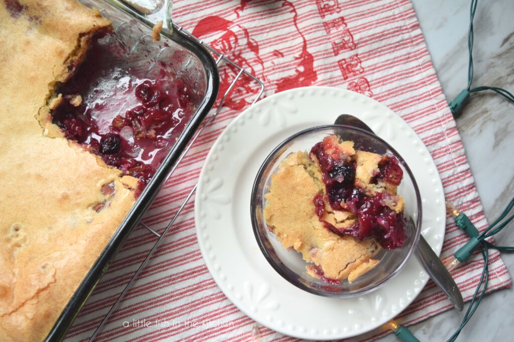 A glass baking dish with one serving of cobbler is shown. The deep red fruit filling is pooled up in the open space and the golden brown cake topping is seen from the top. A single serving of cobbler is in a glass bowl that is on a small white plate. There is a red and white striped tea towl with a picture of Santa Claus and the word "believe" embroidered on to it. Small white lights are scattered around the table. 