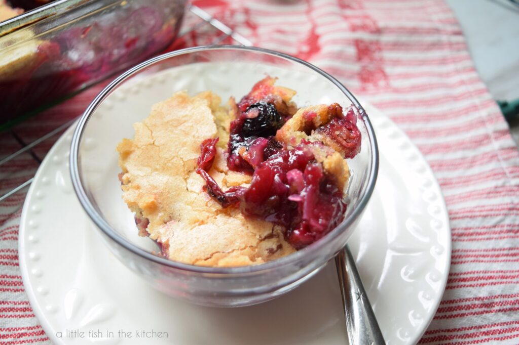 A close up shot of a serving of cranberry cobbler. The golden crust looks crisp and flaky and the dark red fruit filling is visible from the top. The clear glass bowl is on a white plate with a spoon beside it. The plate is on top of a red and white striped tea towel. 