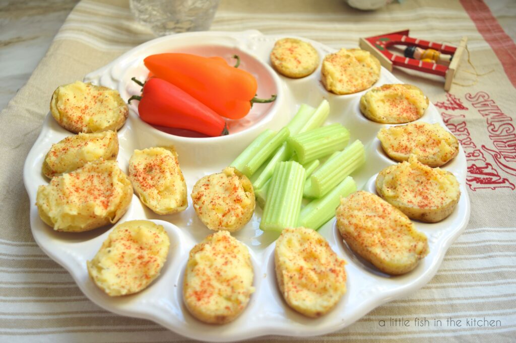 Fourteen deviled potatoes are resting on a deviled egg plate. The plate also has celery sticks and baby bell peppers surrounding it. The plate is on a holiday theme tea town that is beige with with stripes. At the bottom of the towel is red embroidered Christmas tree and the words "Merry Christmas. A small Christmas ornament with a soldier is is laying on the corner of the towel. 