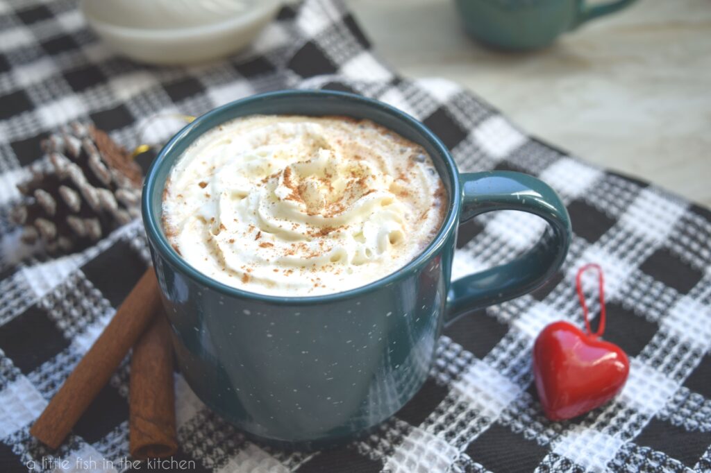 A close up shot of a dark teal coffee mug is filled with Cinnamon Mocha Latte and topped with swirl of whipped cream sits atop a black and white checkered tea towel. Two cinnamon sticks and a red, heart-shaped ornament sit beside the mug. 