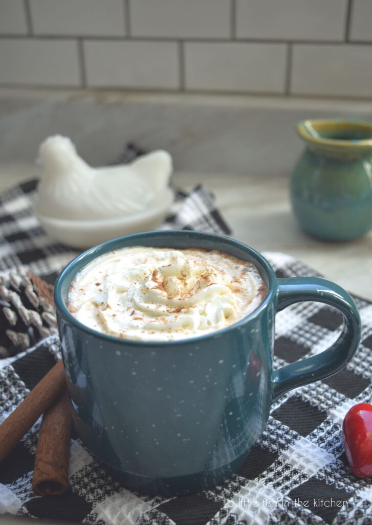 A close up shot in a portrait style photo of a dark teal coffee mug  filled with Cinnamon Mocha Latte and topped with swirl of whipped cream sits atop a black and white checkered tea towel. Two cinnamon sticks and a red, heart-shaped ornament sit beside the mug. A decorative ceramic, chicken and a small, teal ceramic mug are slightly blurred in the background against a white subway tile wall. 