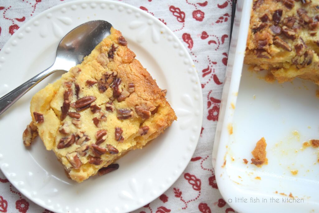 A square slice of cake sits on top of a white dessert plate. The top of the cake is golden and several pieces of chopped pecan are on top. The white baking dish with the whole cake sits next to the plate and a white towel with a red floral pattern is in the background.