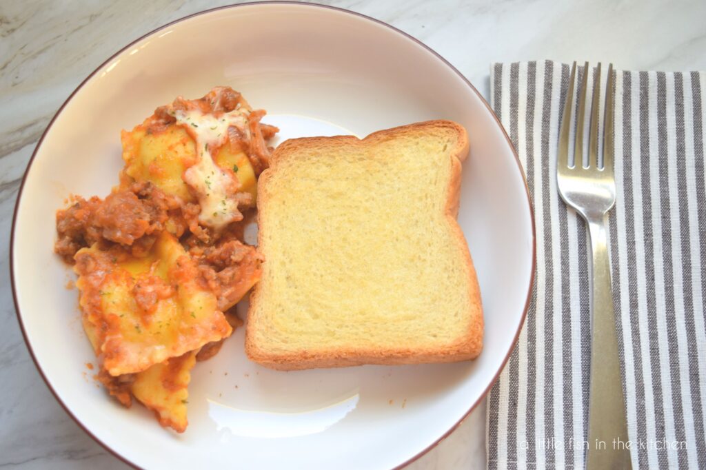 A single serving of ravioli with meat sauce and melted cheese along with a piece of garlic toast. The meal is served on a small white plate and a graw and white cloth napkin is on the side with a fork on top. 